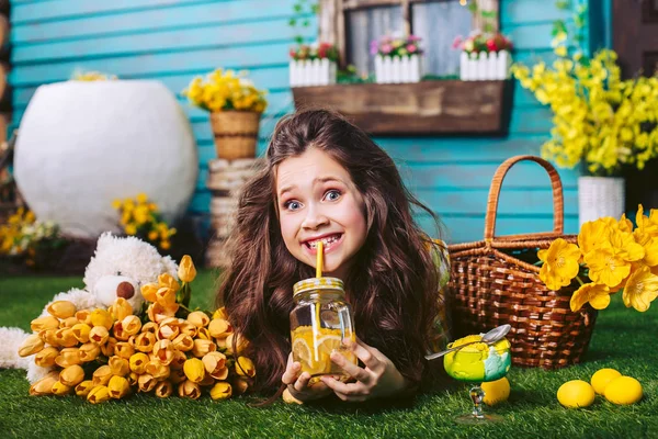 Uma menina brincalhona faz rostos deitados em um gramado perto da casa cercada por flores amarelas. Uma criança bebe refrigerante e come sorvete . — Fotografia de Stock