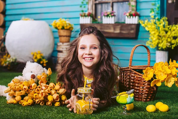 Piękna dziewczynka otoczona dużą liczbą żółtych kwiatów leży na zielonym trawniku w pobliżu drewnianego domu. Dziewczyna napojów lemoniady i zjada lody. Święto wielkanocne. — Zdjęcie stockowe