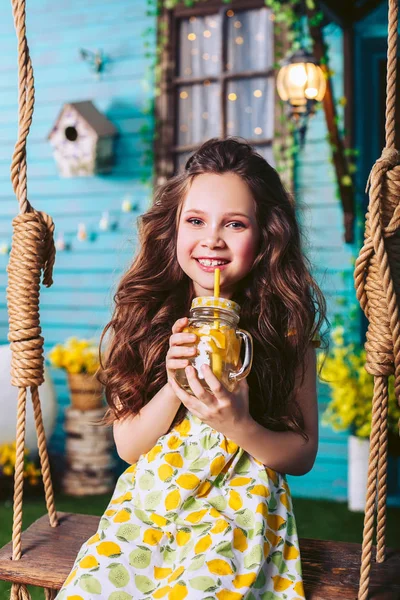 Chica morena en un hermoso vestido sentado en un columpio en el pueblo bebiendo limonada . Fotos De Stock Sin Royalties Gratis