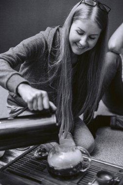 Genç ve güzel bir kız küçük bir masada yerde otururken ve gülümseyerek çay yapıyor.