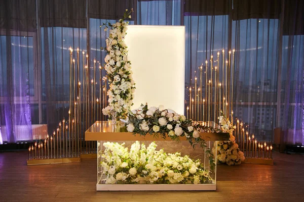 Een glazen bruiloft tabel van de jonggehuwden versierd met een weelderig boeket van natuurlijke bloemen in wit. Garland van metalen buizen en lampen op de achtergrond. Stockfoto