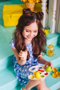 Bir çocuk evin yakınındaki merdivenlerde oturan farklı renklerde boyalar ile Paskalya yumurtaları boyar. Mavi elbiseli kız gülümsüyor..