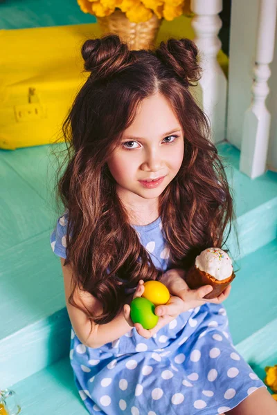 Ein hübsches kleines Mädchen mit dunklen Haaren sitzt auf den Stufen und hält in der Hand einen Osterkuchen und Eier von gelber und grüner Farbe. — Stockfoto