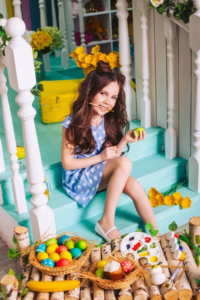 一个女孩坐在台阶上，牙齿上拿着一把刷子给复活节彩蛋着色。一个可爱的孩子庆祝复活节. — 图库照片