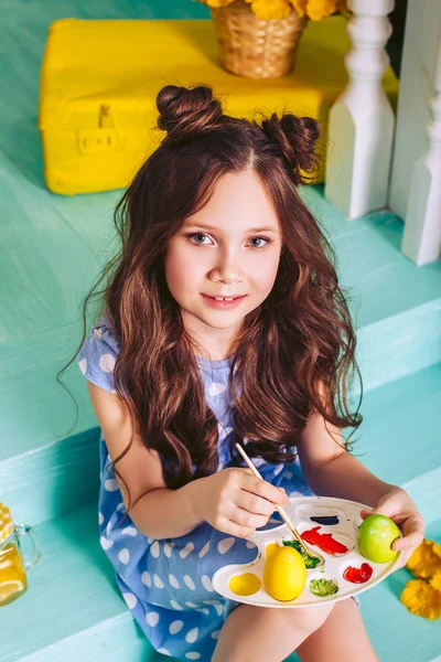 A beautiful little brunette girl paints with colour Easter eggs sitting on the steps of a house in a blue dress. — Stock Photo, Image