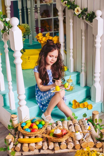 En brunett flicka sitter på trappan i studion och målar påskägg. Förberedelse för ljus påsk. Ägg och påsk kakor i korg korgar på ett bord av trä. — Stockfoto