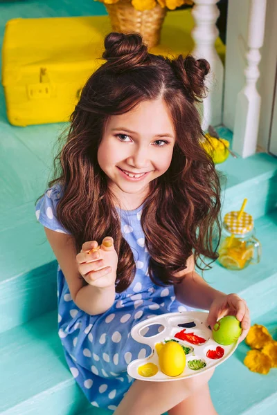 陽気な小さな女の子は、家の階段に座って塗料でイースターエッグをペイントします。ホリデーイースター. — ストック写真