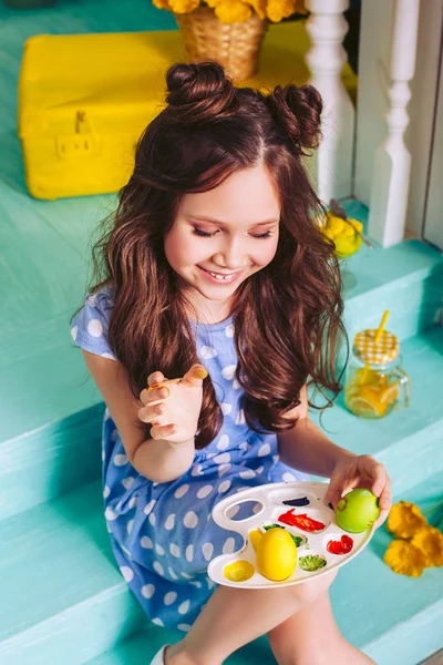 Un enfant peint des œufs de Pâques avec des peintures de différentes couleurs assis sur les marches près de la maison. La fille dans la robe bleue sourit . — Photo