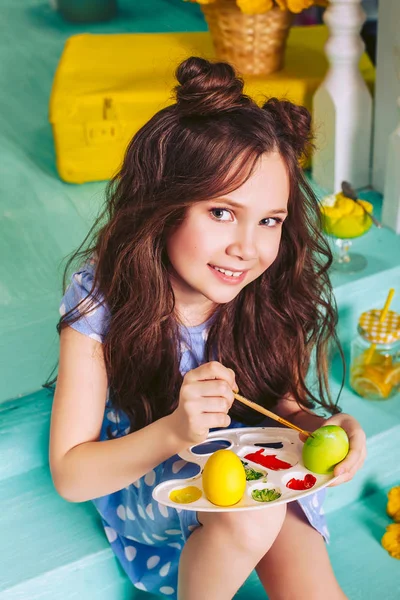 Una linda chica en un vestido azul sentado en los pasos pintados huevos de Pascua con pinturas de colores . — Foto de Stock