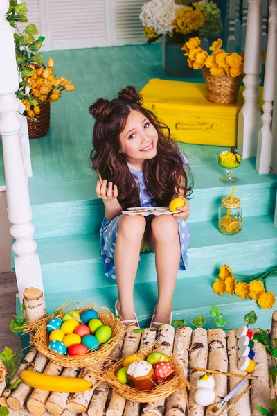 Маленькая девочка позирует, раскрашивая пасхальные яйца, сидя на ступеньках возле дома в красивом голубом платье . — стоковое фото