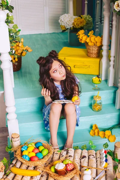 Una ragazza in un abito blu a pois posa il viso e dipinge uova di Pasqua seduta sui gradini della casa . — Foto Stock