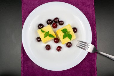 Beyaz bir tabakta iki parça süzme peynir güveci, kirazla süslenmiş. Siyah arkaplan