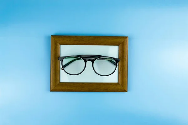 Round glasses in black frames lie in a photo frame. Blue background