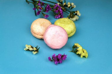 Mavi arka planda çok renkli badem kurabiyeleri, çok renkli çiçeklerin yanında.