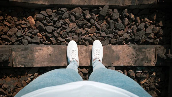 Female legs in jeans and white sneakers on railroad tracks from the first person