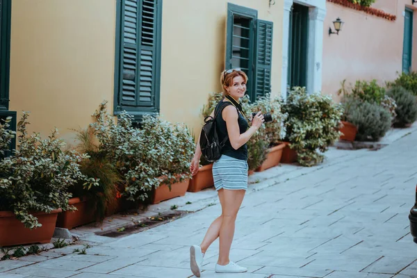 Hermosa chica caminando por la calle de un viejo pueblo europeo con una cámara de cine en la mano. Retrato de una chica turística caminando sobre el fondo de la calle. — Foto de Stock