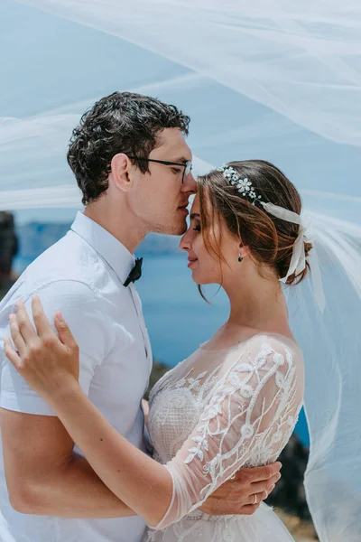 Прекрасна наречена обіймає і цілує під весільною вуаллю в день їхнього літнього весілля на острові Санторіні. — стокове фото