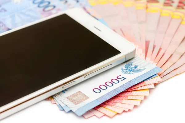 Smartphone above the piles of Indonesia Rupiah (IDR) banknotes on the white isolated background.