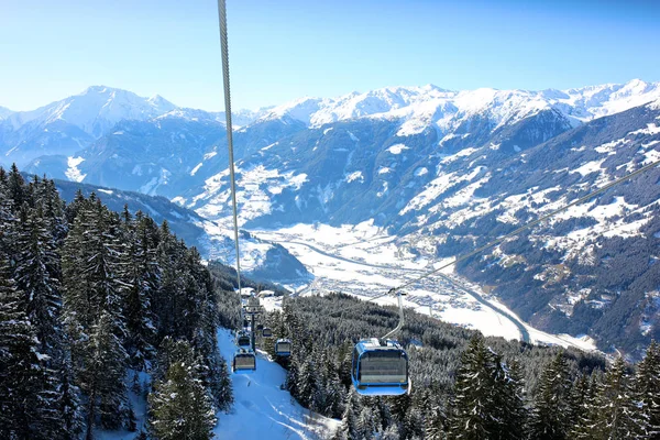 Modern comfortable gondola lift in Alpine ski resort. People enjoying skiing on prepared slopes in the Alps on sunny day. Beautiful snowy trees and little village in the mountains. Perfect winter holidays destination for family.