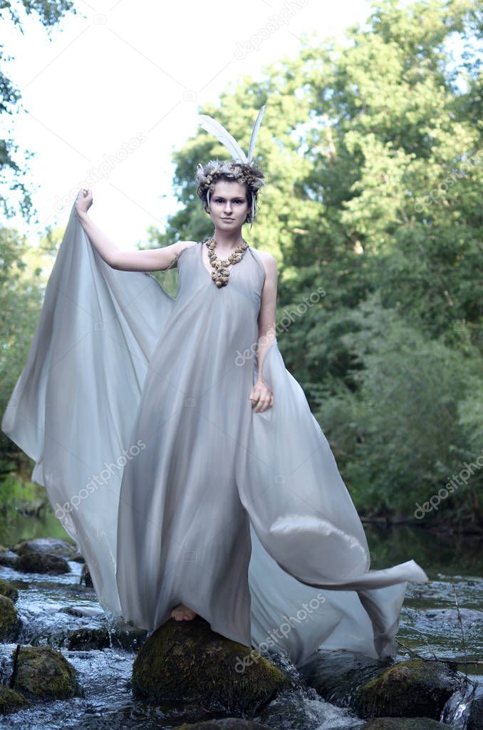 young woman in a gray silk dress, a headdress of feathers and a necklace of sea snail shells stands on the rocks among the flow of a stormy river and waves the train of her dress