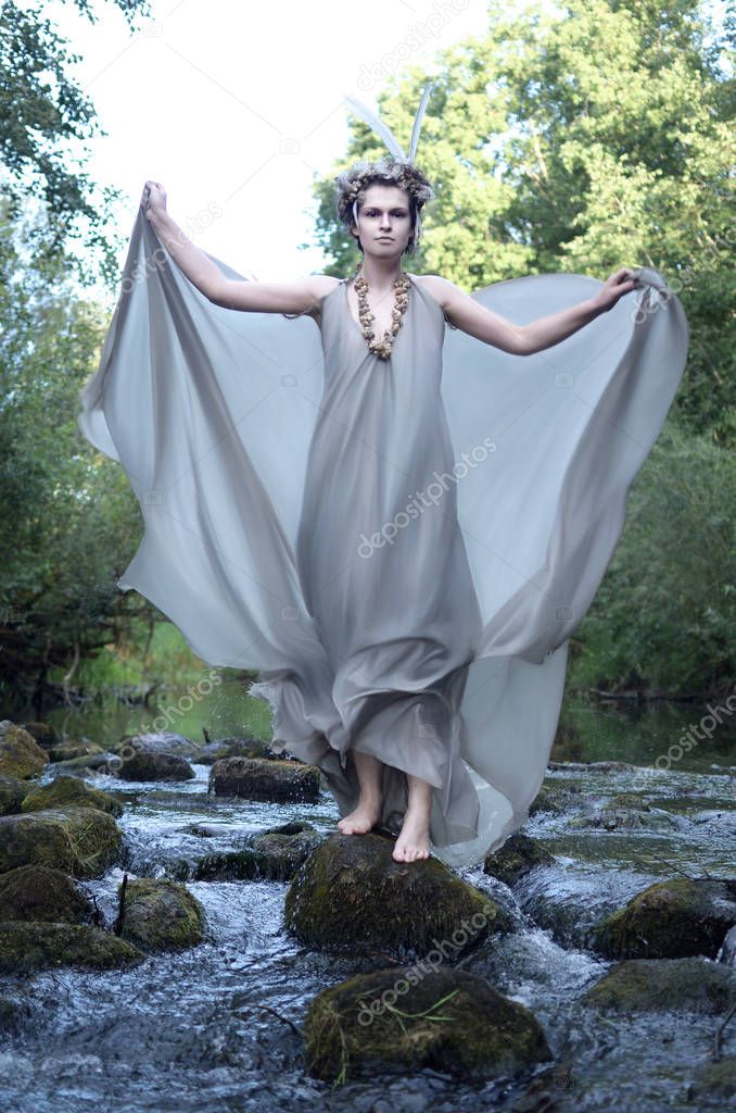 young woman in a gray silk dress, a headdress of feathers and a necklace of sea snail shells stands on the rocks among the flow of a stormy river and waves the train of her dress