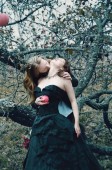 junger Mann mit langen blonden Haaren küsst ein dunkelhaariges Mädchen in den Nacken unter einem fliegenden Apfelbaum in einem alten Garten