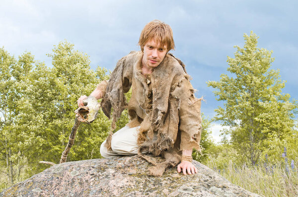 young shaman with a stick from an old stick sits on a huge stone and looks at something with interest