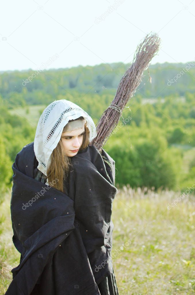 young witch in a black cloak and dress stands with a magical broom on top of a mountain on a Sabbath