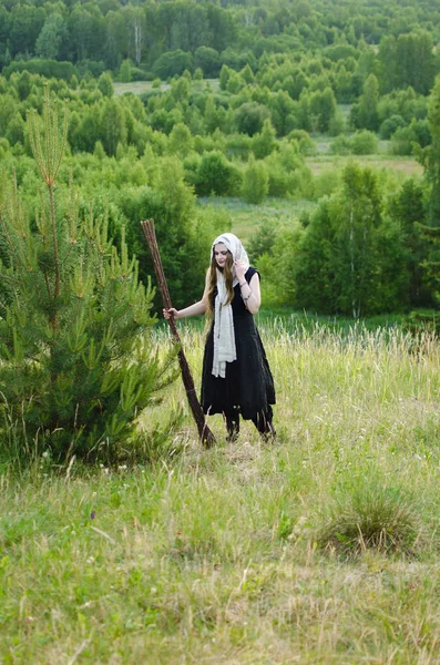 witch with a broom and a black dress stands in the woods on a grassy hill