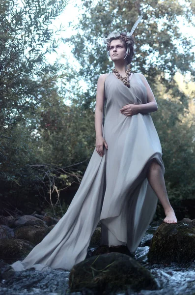 güzel ve genç birbayan gri ipek elbise, tüy headdress ve deniz salyangozları orman perisi, dere taşı üzerinde duran ve dinlenme bir kolye