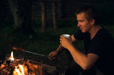 adam oturur ve şömine başında bir şiş üzerinde bir sosis kızartma aittir