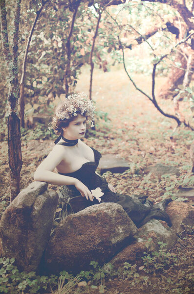 Young woman in a lush black dress sits in an old garden amidst a pile of large stones