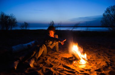 young tourist woman pulls her hand to the night fire on the beach clipart