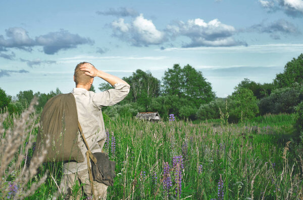солдат в старой военной форме Второй мировой войны в поле среди высоких диких трав с большим мешком в руках, кладет руку за голову, глядя вперед
