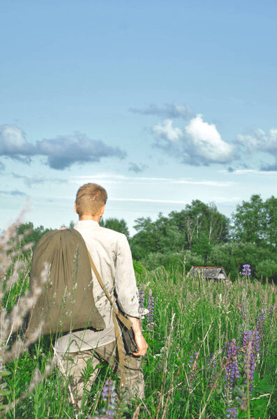 человек в старой форме Второй мировой войны в поле среди высоких диких трав с большим мешком в руках

