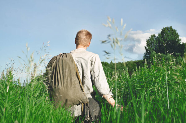 Человек в старой военной форме на поле трогает траву руками и тащит большую сумку.
