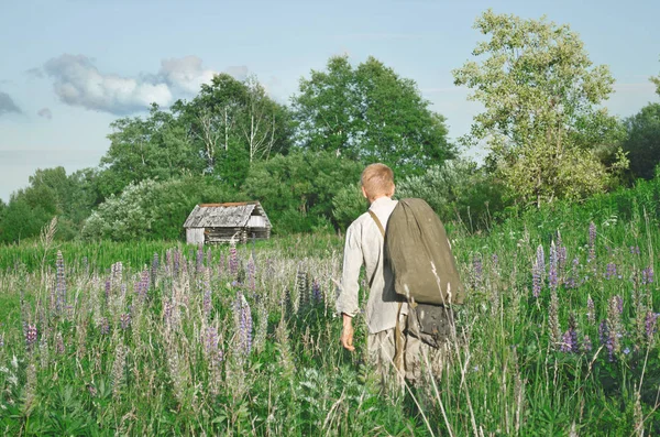 一个拿着包的人在田野里的高大的草中间 望着旁边 在远处你可以看到一座破旧的村屋 — 图库照片