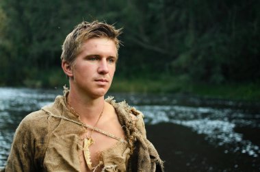 portrait of a young medieval peasant man in a shirt of old burlap on the background of the forest and the river clipart