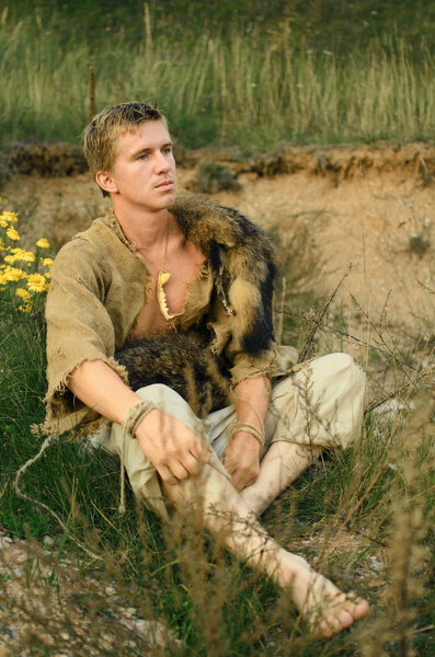 портрет молодого средневекового крестьянина или охотника в старом мешковине с кожей животного на плечах, лежащего в поле
