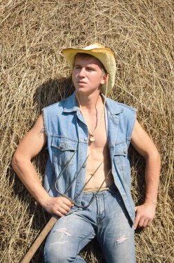 Elinde yabayla genç ve seksi bir adam bir çiftlikte samanlığın yanında duruyor.