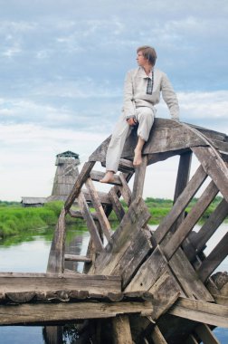 young man in a national costume of white linen sits on an old water mill wheel and looks into the distance clipart