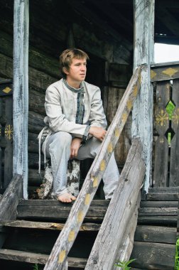 young man in old linen clothes is sitting on the wooden porch of a village hut clipart