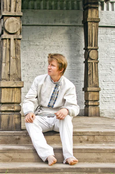 young man in old linen clothes is sitting on the steps of the porch of a wooden Terem