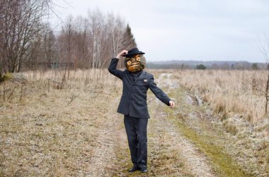 Kafasında bir cadılar bayramı balkabağı olan korkunç korkuluk kırsal bir yolda duruyor takım elbise giymiş ve yoldan geçenleri selamlayarak onları korkutuyor.