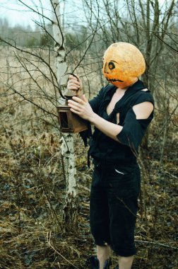 Kafasında bir balkabağı ve ellerinde antik bir fenerle Cadılar Bayramı Korkuluğu karanlık bir sonbahar gününde ormanda dolaşır.