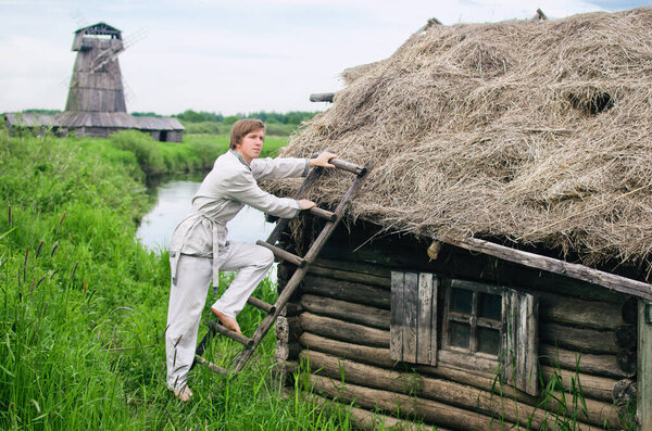 village young man in white linen clothes climbs up a ladder to the thatched roof of a small hut