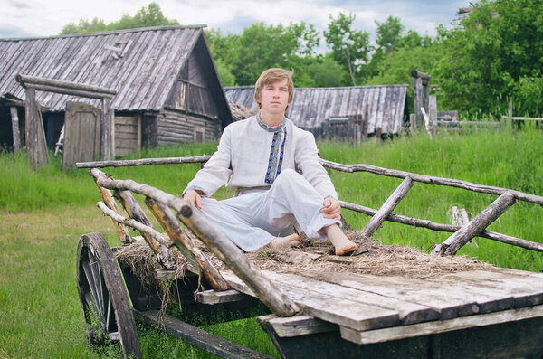 young man in old linen clothes is sitting in an old cart in the village