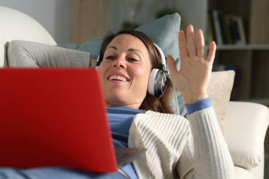 Happy adult woman videocalling with headphones waving on laptop lying on a sofa in the livingroom at home clipart