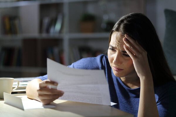 Worried girl reads bad news on letter at night sitting in the living room at home