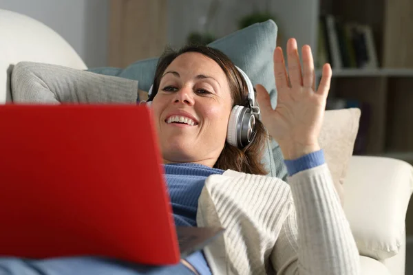幸せな大人の女性ビデオコーリングともにヘッドフォンを振ってラップトップ上に横たわる上のソファの上に自宅のリビングルーム — ストック写真